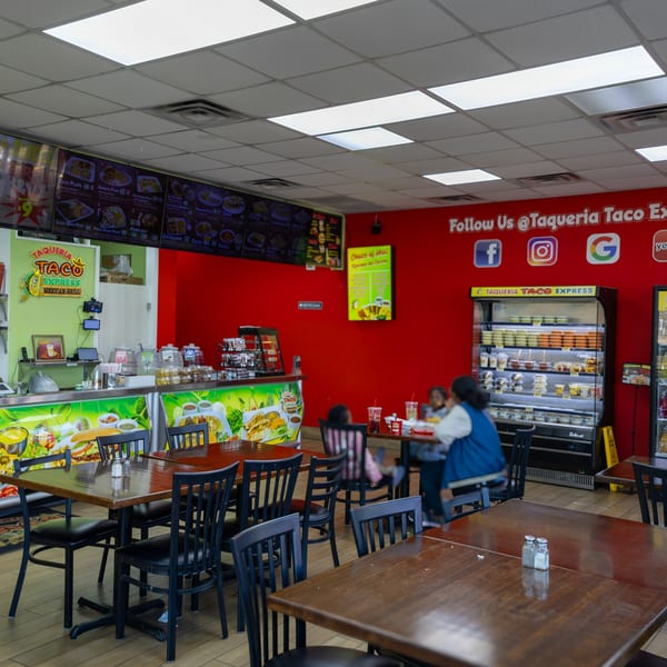 Interior of Taqueria Taco Express, showing dining tables, a counter, and a red wall with social media logos.