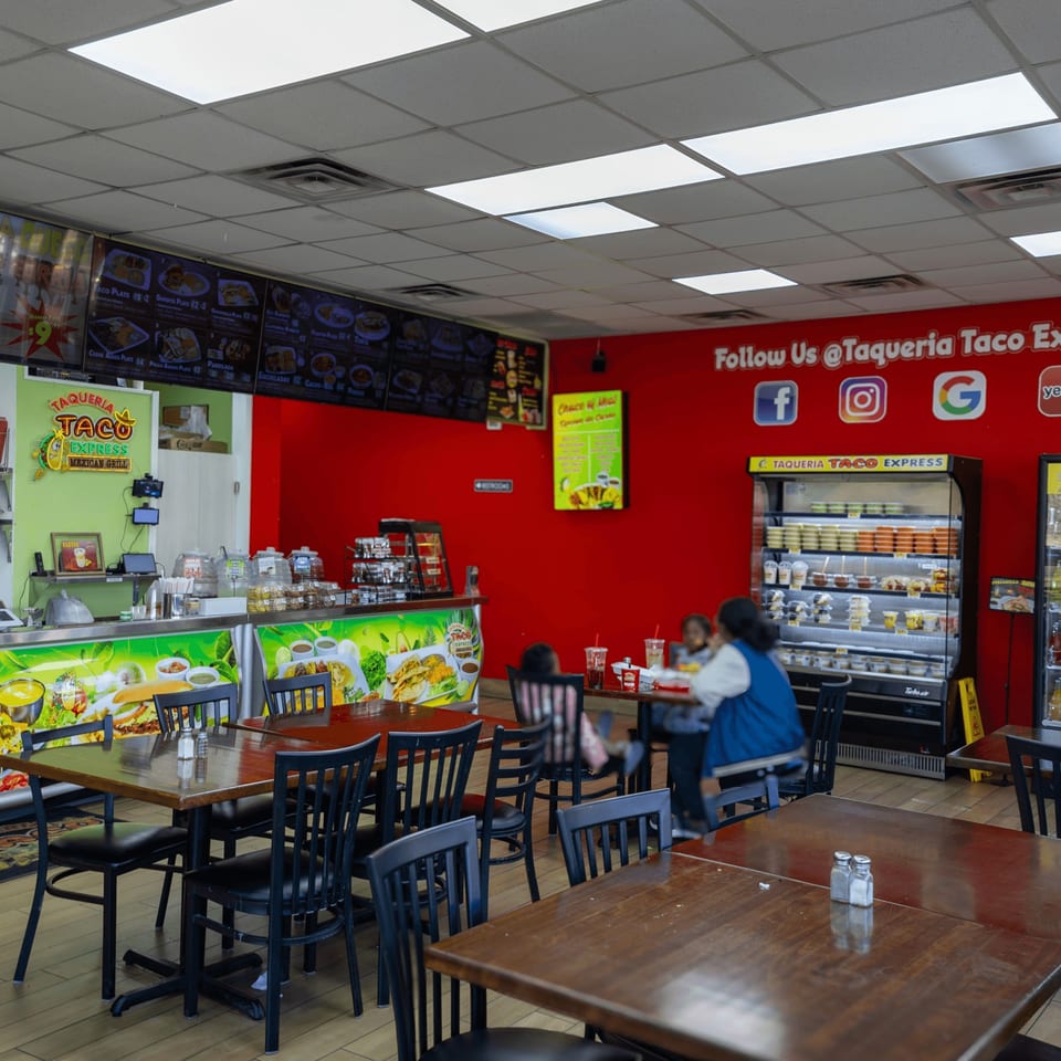 Interior of Taqueria Taco Express, with red walls, menu boards, a counter, and dining tables with customers.