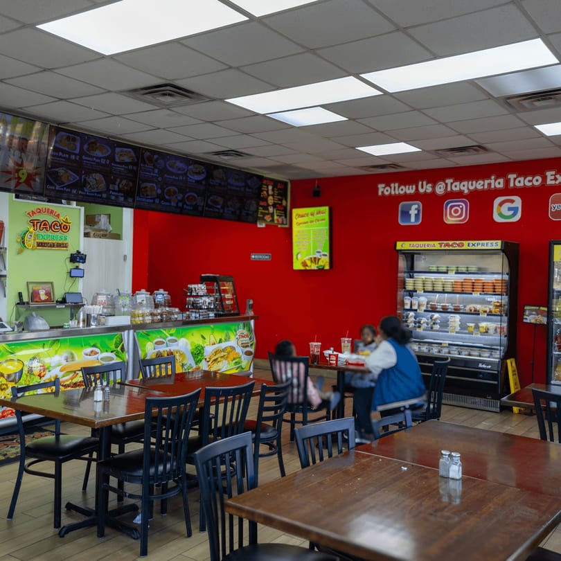 Interior of Taqueria Taco Express, with red walls, menu boards, a counter, and dining tables with customers.