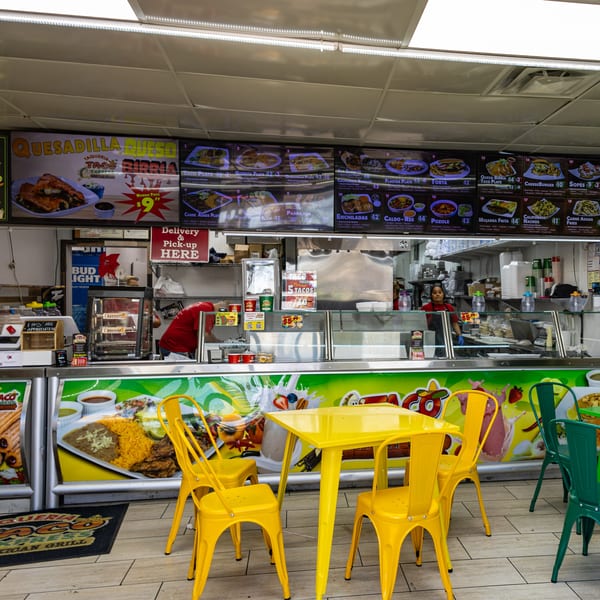 Interior of Taqueria Taco Express with menu boards, food prep area, and yellow and green tables.